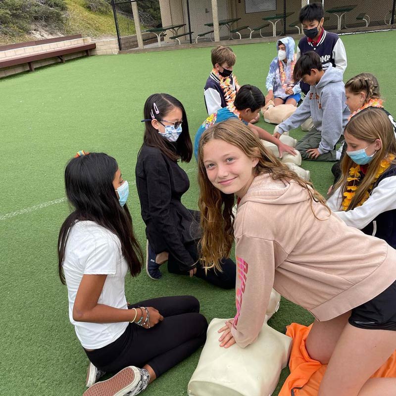 A group of Del Mar Pines students practice CPR on mannequins outdoors on a grassy field.