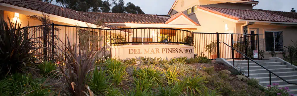 A view of Del Mar Pines School’s entrance, featuring a beige building with a red tile roof, a lighted sign, black metal fencing, steps, and landscaped greenery.