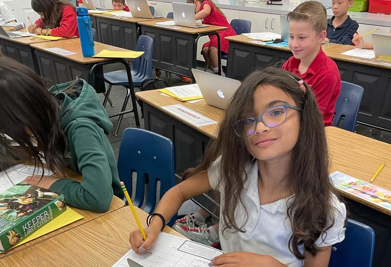 A girl with glasses sits at a classroom desk, smiling and writing on a worksheet. Other students are behind her, working on laptops. The classroom is bright and organized.