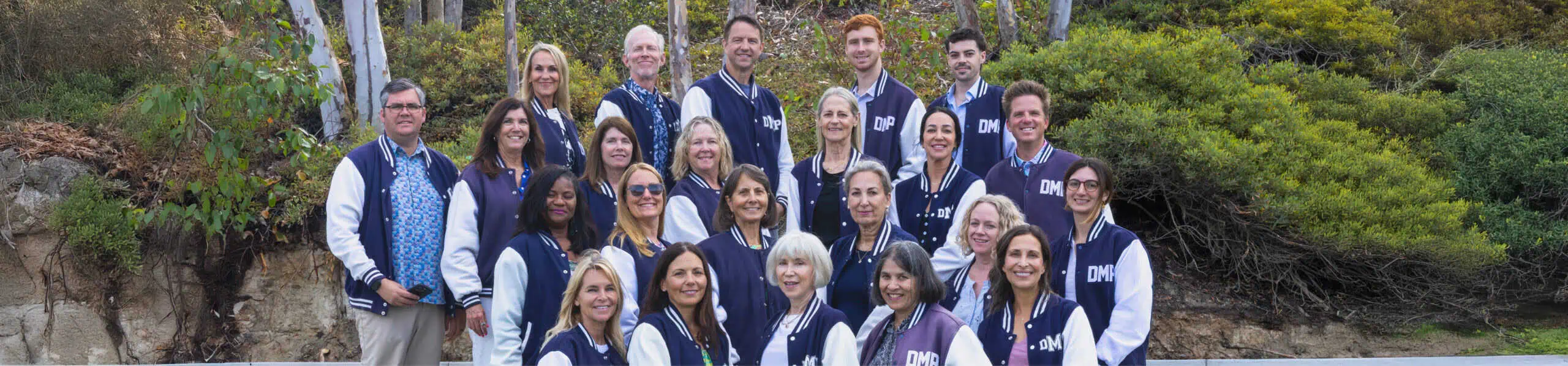 DMP teachers wearing matching navy and white jackets posing outdoors in front of greenery and trees, smiling at the camera.