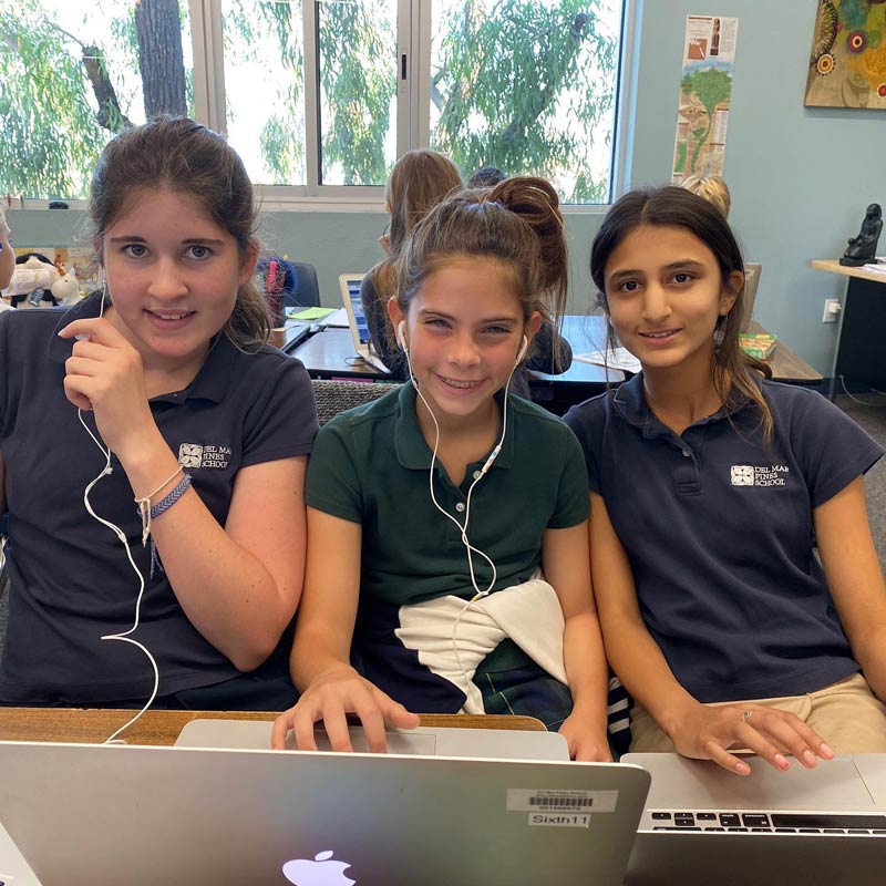 Three Del Mar Pines School girls sit together at a table in a classroom, wearing earphones and smiling at the camera. Two of them are using a laptop, and there are other students and classroom materials in the background.