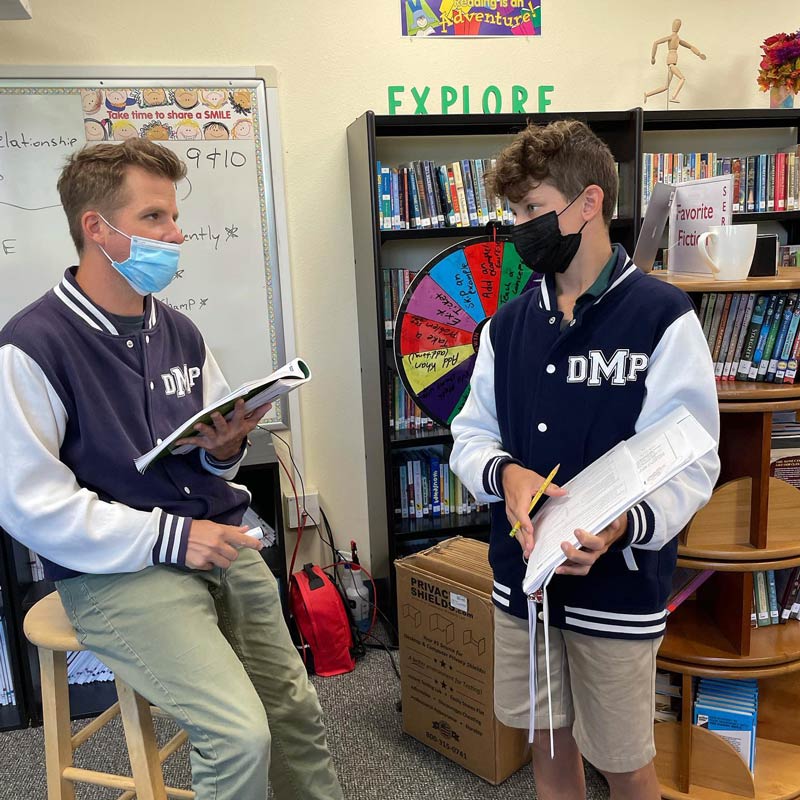 A Del Mar Pines School teacher and student wearing face masks and matching jackets talk in a library.
