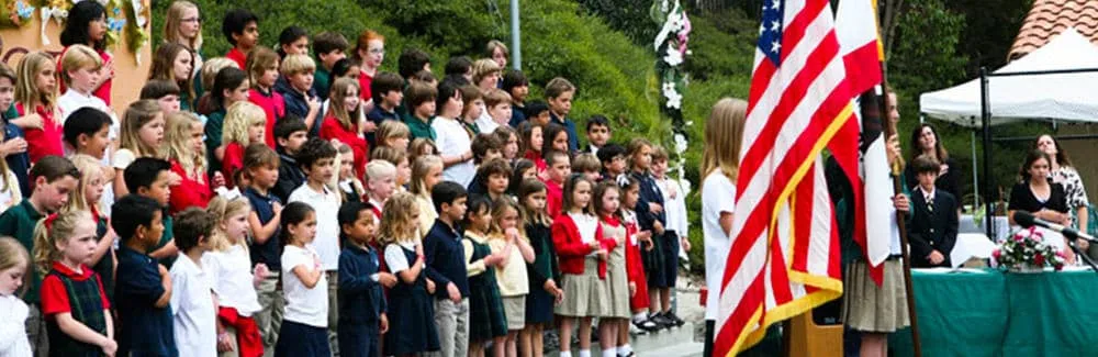 A large group of Del Mar Pines school children stand outdoors, facing the American flag during a ceremony.