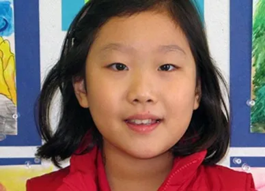 A young girl wearing a black face mask stands in a classroom, holding up a Eureka Math Grade 4 Modules 1 & 2 book. Classroom decorations and cabinets are visible in the background.