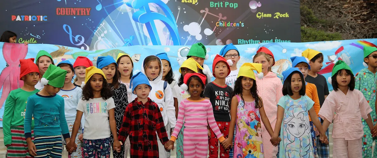 A diverse group of children stand in a row, wearing colorful hats and casual clothes, holding hands and singing on stage in front of a bright, music-themed backdrop.