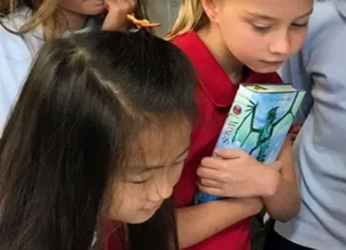 A young girl wearing a black face mask stands in a classroom, holding up a Eureka Math Grade 4 Modules 1 & 2 book. Classroom decorations and cabinets are visible in the background.
