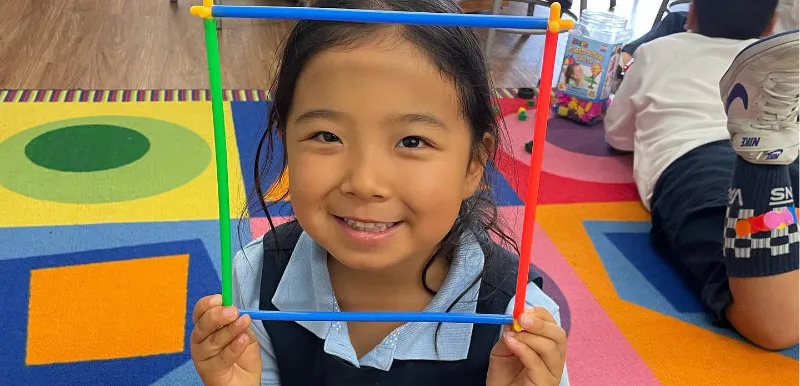 A young girl wearing a black face mask stands in a classroom, holding up a Eureka Math Grade 4 Modules 1 & 2 book. Classroom decorations and cabinets are visible in the background.