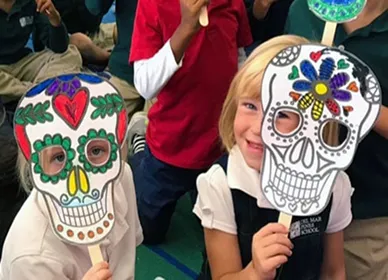 A young girl wearing a black face mask stands in a classroom, holding up a Eureka Math Grade 4 Modules 1 & 2 book. Classroom decorations and cabinets are visible in the background.