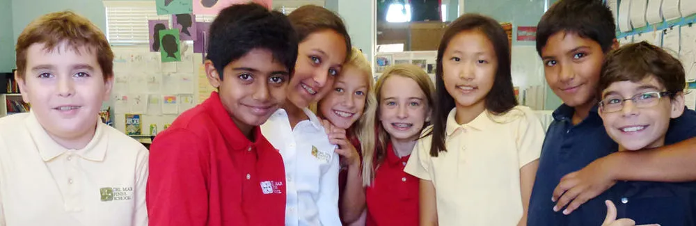 Eight smiling children in a classroom stand close together. Artwork and papers are visible on the bulletin board behind them.